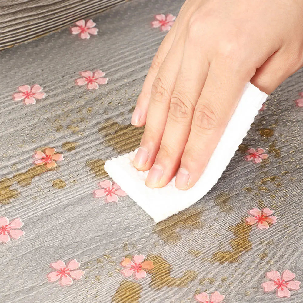 A hand is wiping a surface with a white reusable fridge and cabinet liner pad on a gray textured background.