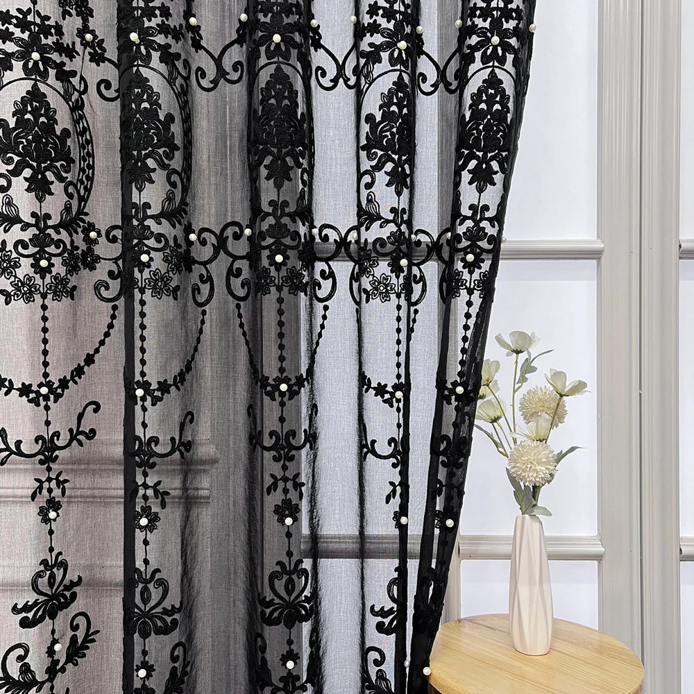 Black sheer curtain featuring intricate lace embroidery with pearl accents hanging in a window.