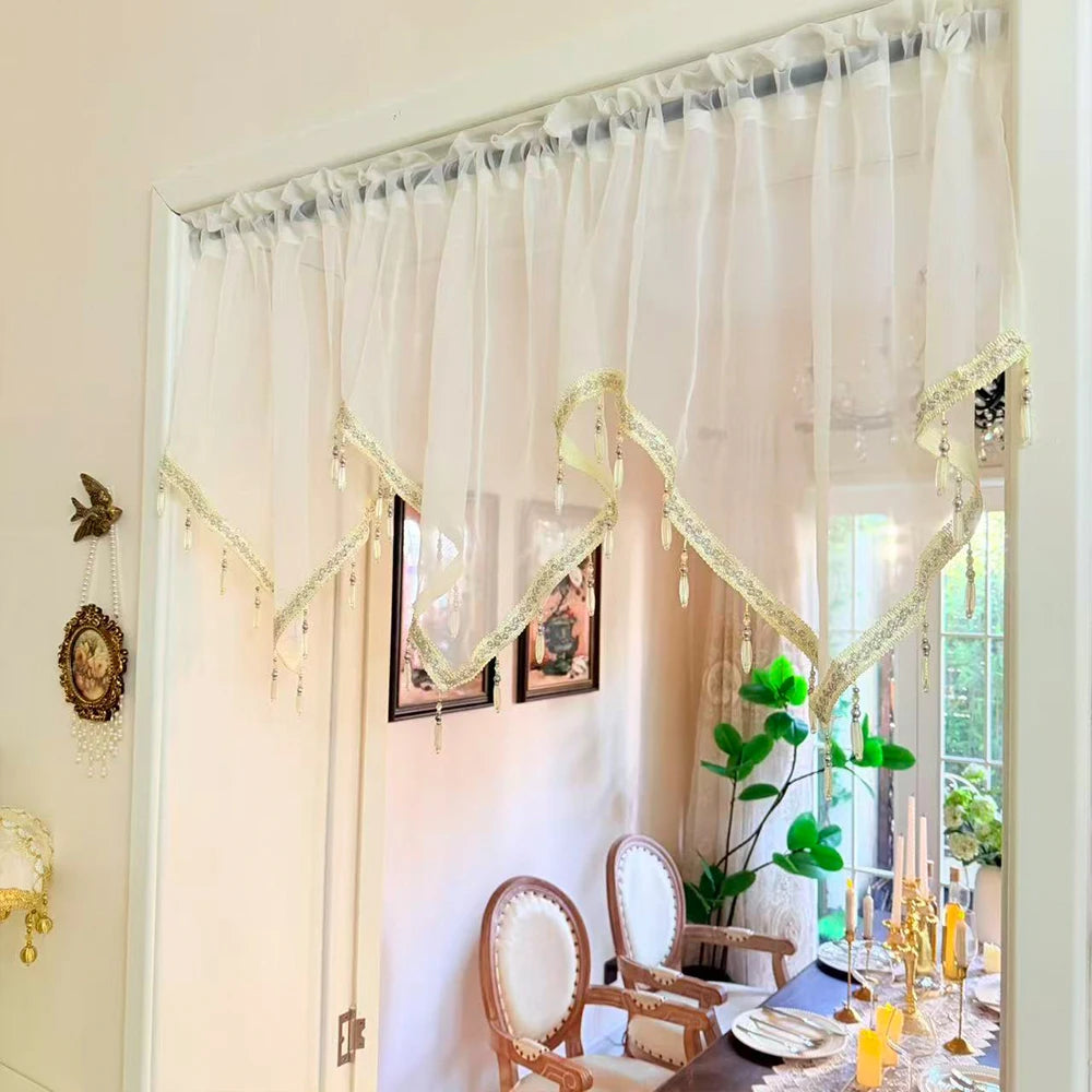 A sheer, ivory curtain adorned with gold lace and tassels hangs in a bright room.
