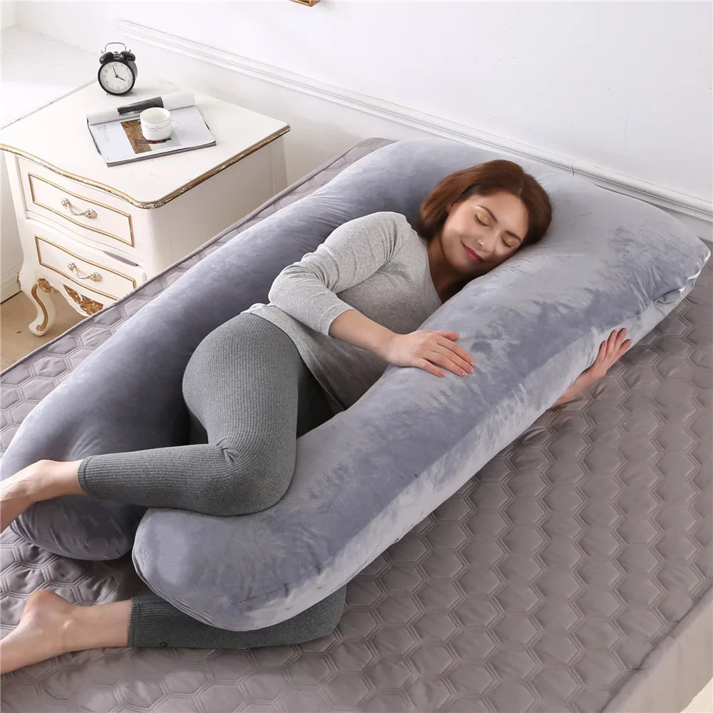 A woman lies comfortably on a gray U-shaped maternity body pillow on a floor.