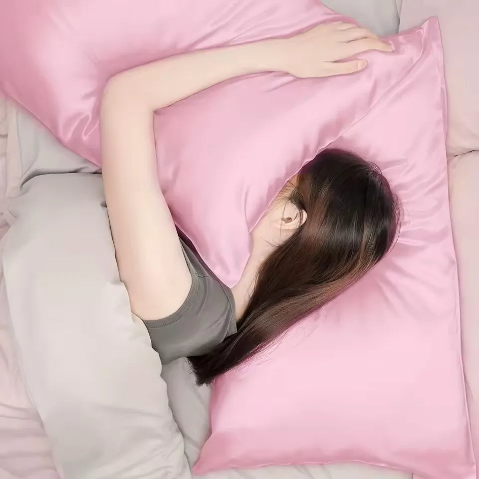 A person with long dark hair is lying down, resting their arm on a pink satin pillowcase.