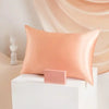 A soft peach colored silk pillowcase resting on a white block, set against a draped fabric background.