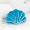 A teal velvet stuffed sofa cushion shaped like a decorative seashell resting on white fabric.