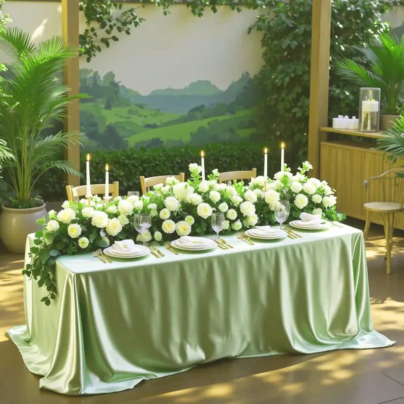 A long table set with white flowers and green satin overlay cloth in an outdoor garden setting.