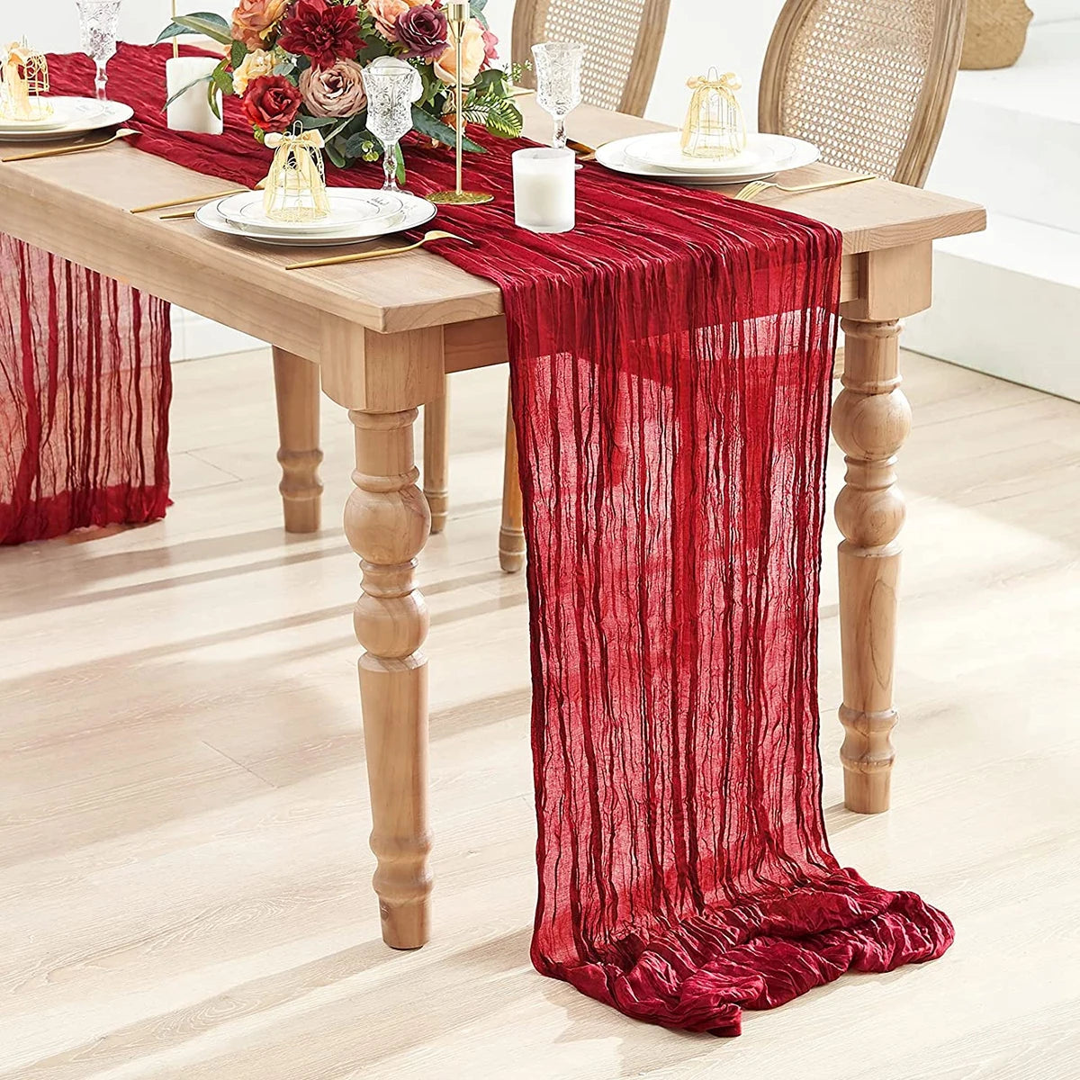 A deep red semi-sheer gauze table runner draped elegantly on a wooden dining table set for a meal.