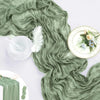 A flowing sage green cheesecloth table runner draped elegantly with white feathers and floral accents.