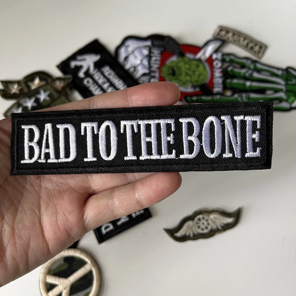A hand holds a black embroidered patch with the text BAD TO THE BONE against a white background.