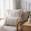 A cream-colored textured throw pillow cover with beige tassels resting on a light gray upholstered chair.