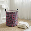 A purple and pink leopard print foldable laundry basket sitting on a woven rug near a window.