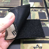 A hand holds a black tactical flag patch against a camouflage fabric background.