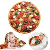 A pizza is shown with a baby lying on a blanket throw featuring pizza toppings.
