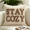 A beige knit pillow cover with the words STAY COZY embroidered in brown yarn on a white sofa.