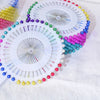 A collection of pearl head pins in various colors and sizes arranged on a white surface.