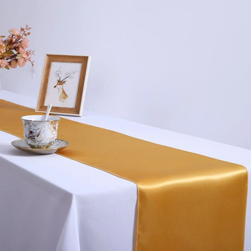 A gold satin table runner draped across a white tablecloth with a framed picture and a small teacup.