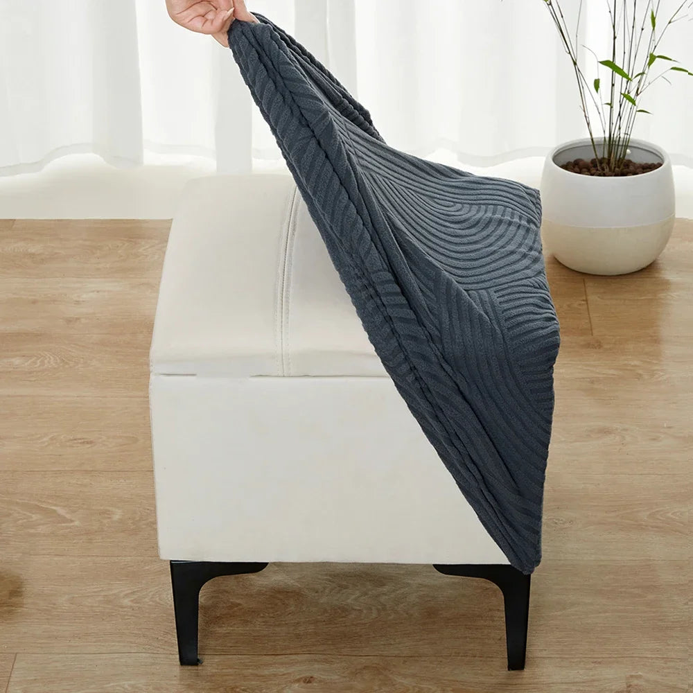 A dark gray geometric jacquard ottoman slipcover draped over a white upholstered ottoman.