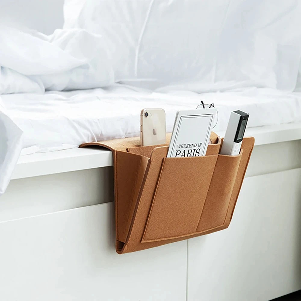 A brown felt storage organizer with hanging pockets holding several smartphones and cards on a white bedside table.