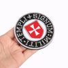 Embroidered military cross shield hook and loop patches on a black circular fabric patch being held by a hand.