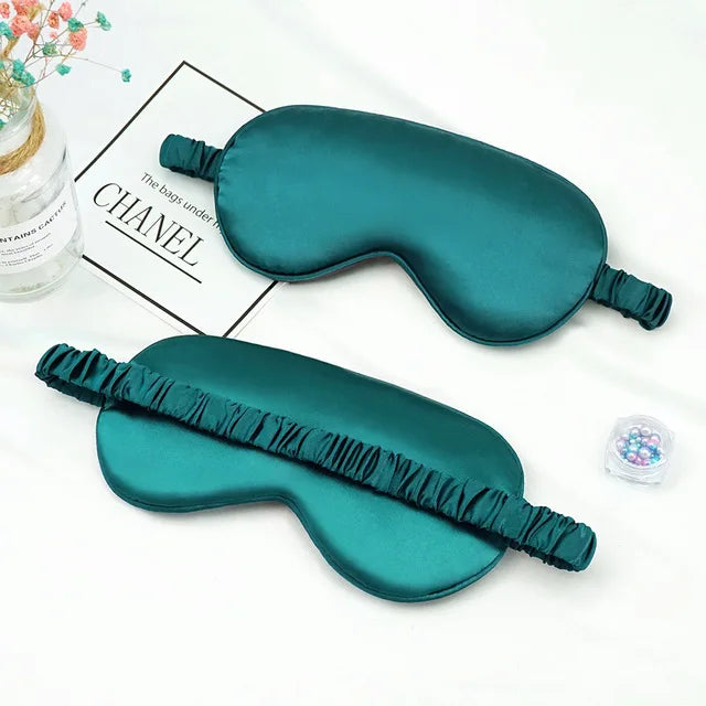 A teal silk sleep mask with a ruffled edge is displayed on a white background next to a Chanel branded box.
