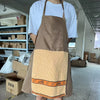 A brown apron with a beige pocket and orange trim is displayed against an industrial background.