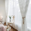 White sheer curtains with floral embroidery hanging in a bright room next to a wooden chair.