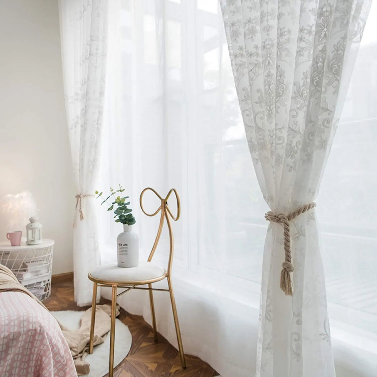 White sheer curtains with floral embroidery hanging in a bright room next to a wooden chair.