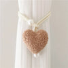 A beige plush heart tieback with a white cord and pearl accents, displayed against a white curtain.