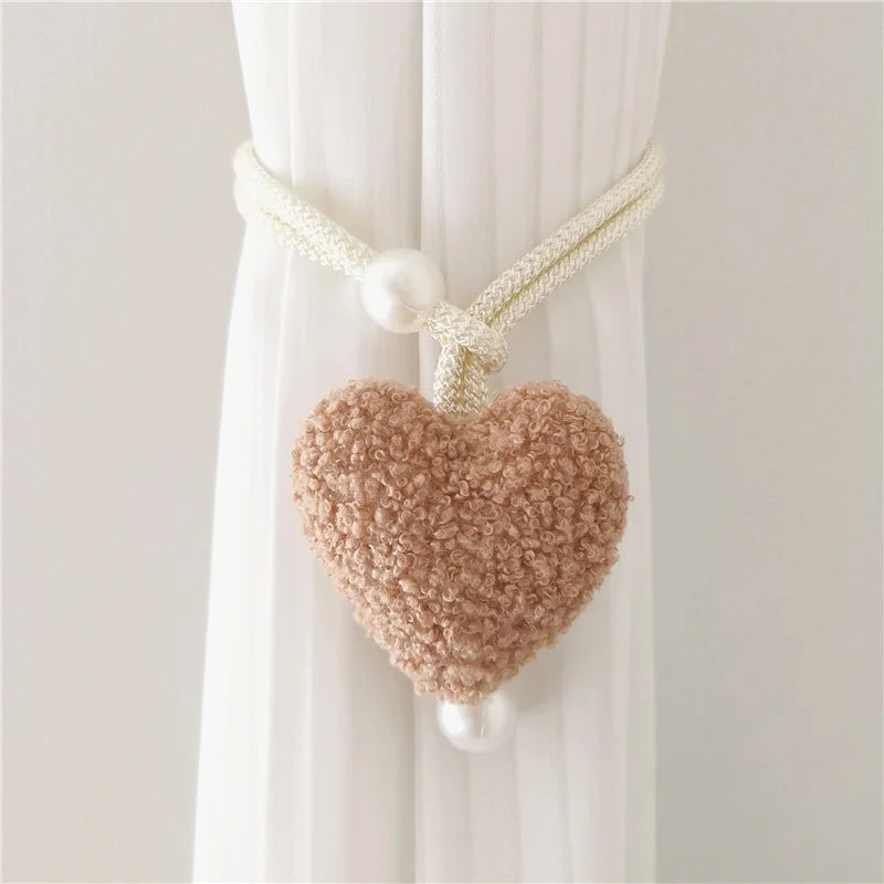 A beige plush heart tieback with a white cord and pearl accents, displayed against a white curtain.