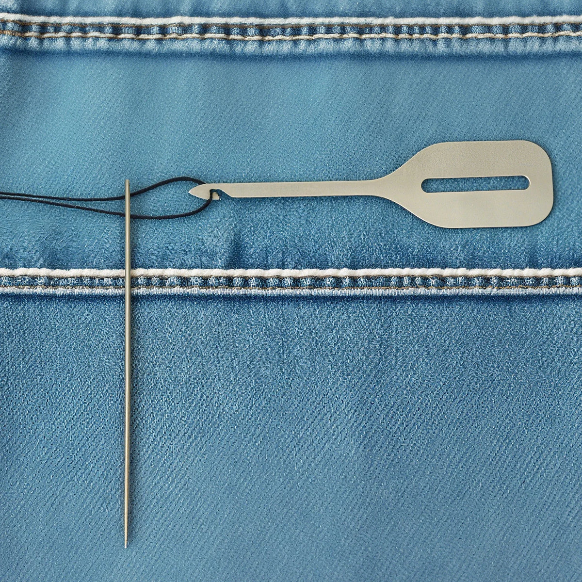 A stainless steel needle threader is shown against a blue denim fabric with stitching.