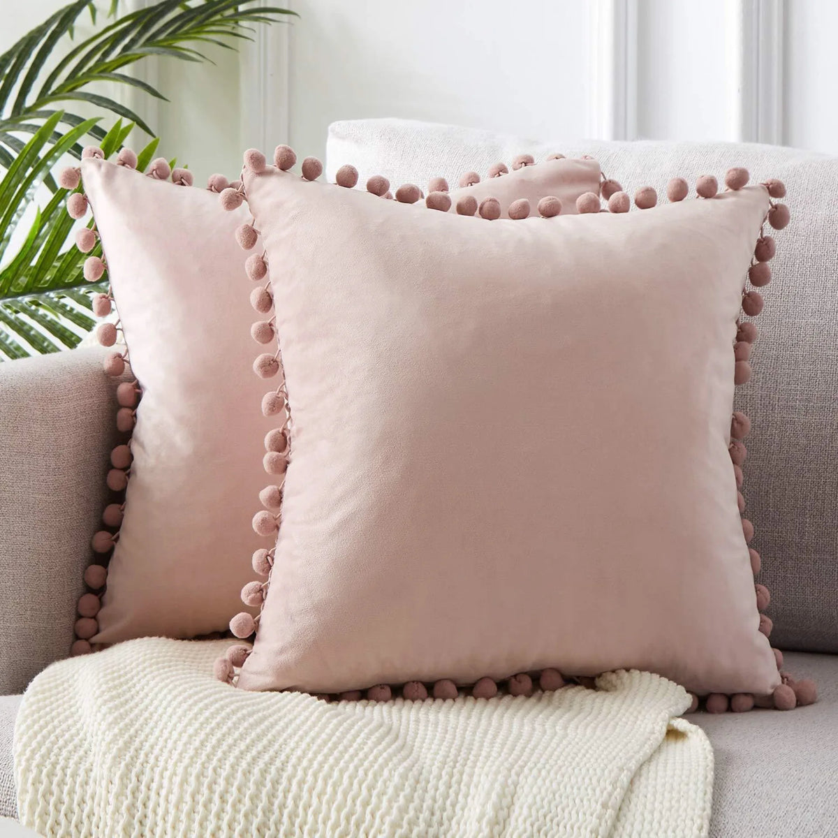 A square, dusty rose velvet cushion cover with decorative buttons rests on a light beige knitted throw.