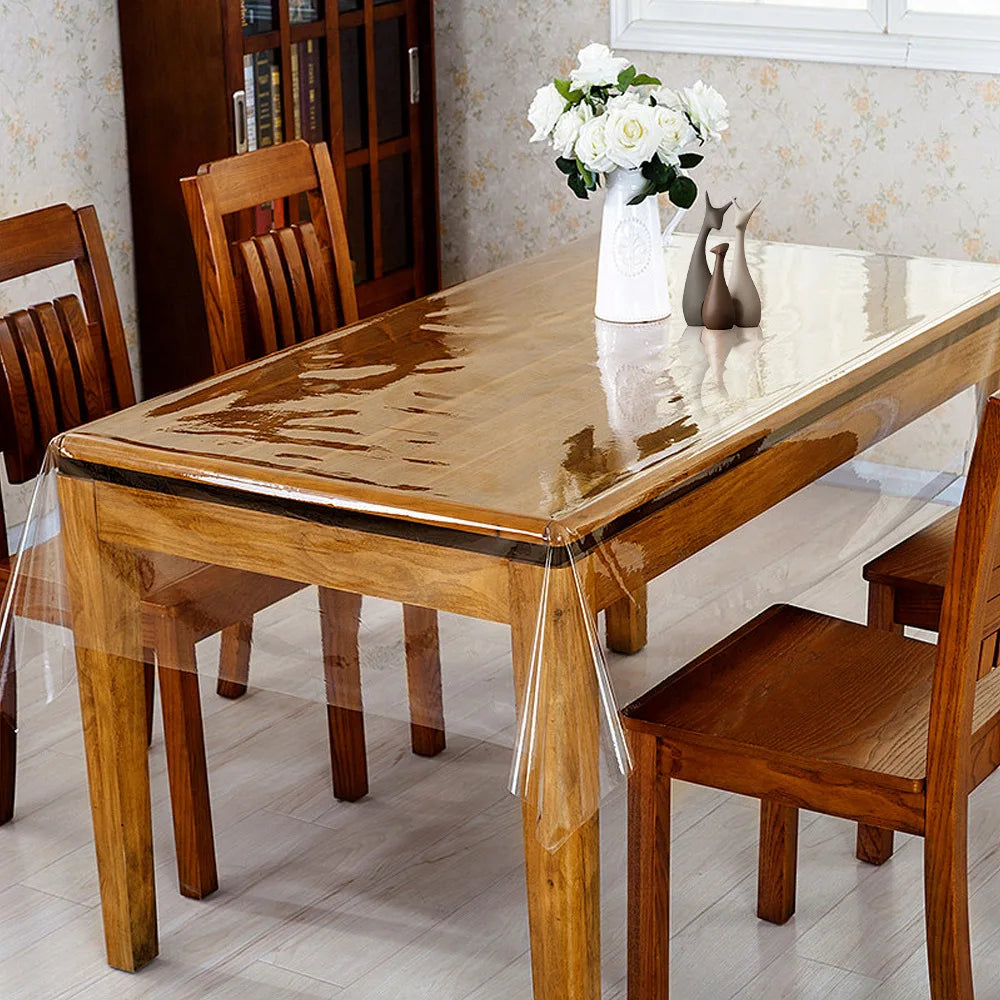 A wooden dining table with a transparent PVC protector mat and wooden chairs, set in a bright room.