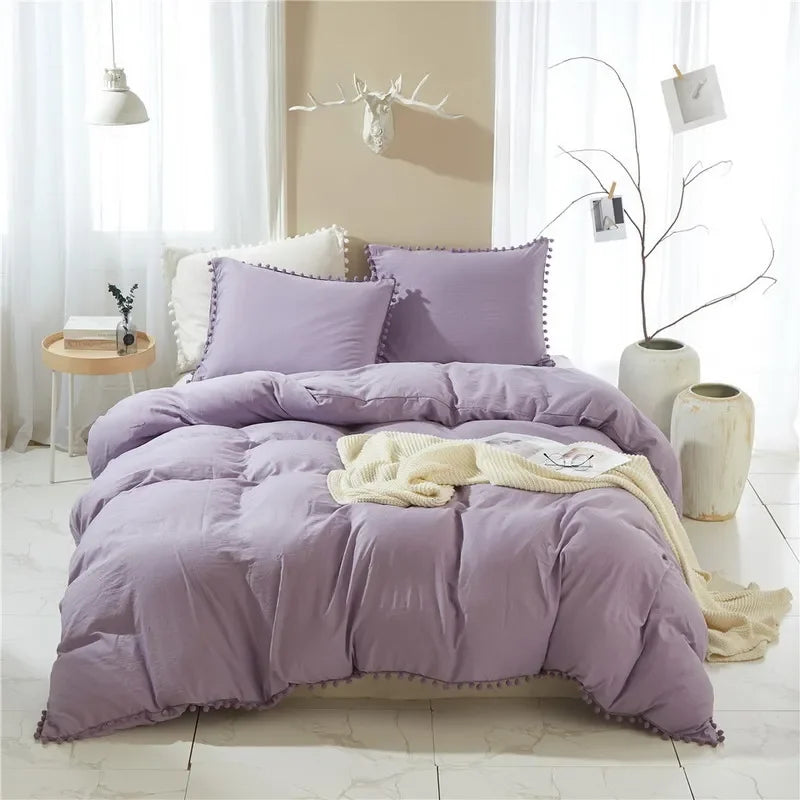A purple quilted duvet cover set with pom-pom decoration displayed on a bed in a bright room.