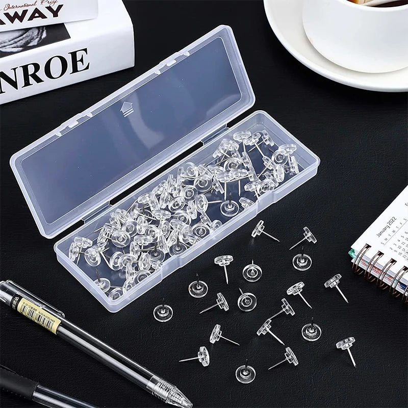 A transparent plastic box filled with round head push pins and a loose pack of similar pins on a dark surface.