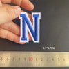 A hand holds a blue embroidered letter N patch next to a ruler showing its dimensions.