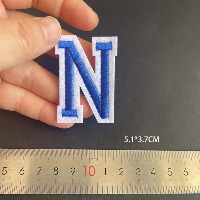 A hand holds a blue embroidered letter N patch next to a ruler showing its dimensions.