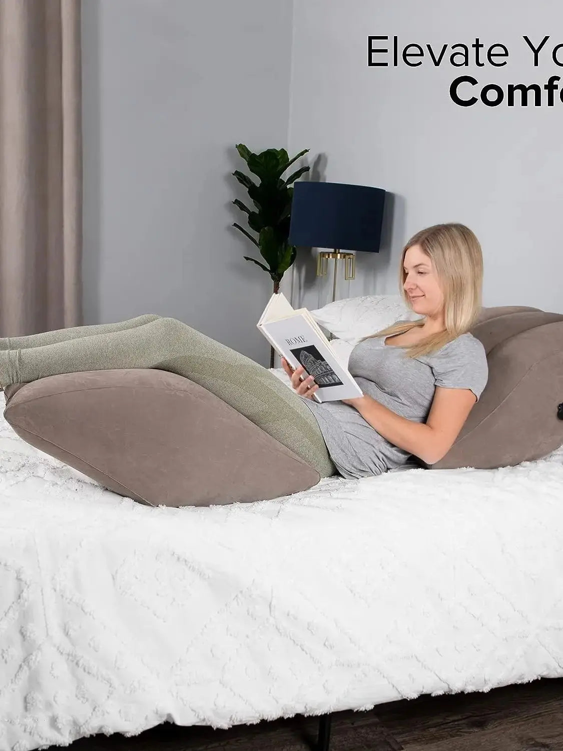 A woman is resting on a bed with an inflatable leg elevation pillow and storage bag, reading a book.
