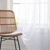A woven rattan chair placed in front of a sheer white tulle curtain by a window.
