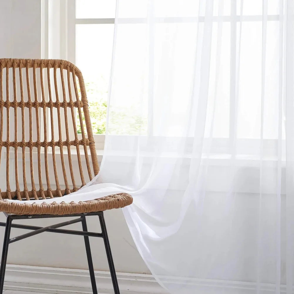 A woven rattan chair placed in front of a sheer white tulle curtain by a window.