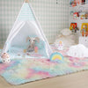 A rainbow plush area rug is displayed on a light wooden floor, surrounded by a white tent and stuffed animals.
