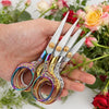 A hand holds a pair of vintage stainless steel sewing scissors with colorful handles against a backdrop of pink and red flowe