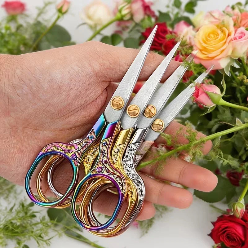A hand holds a pair of vintage stainless steel sewing scissors with colorful handles against a backdrop of pink and red flowe