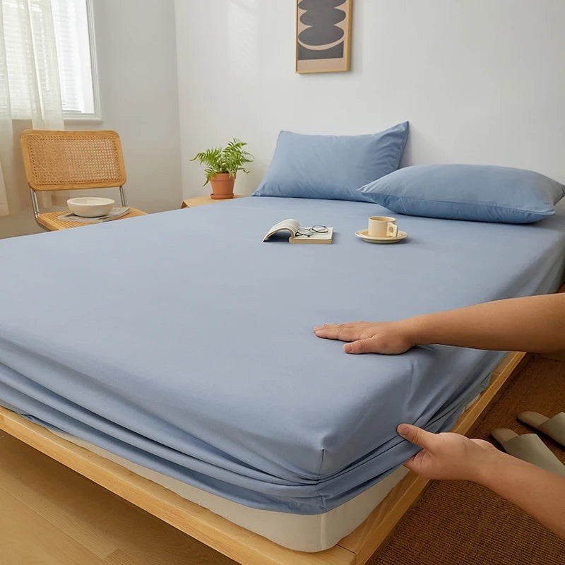 A light blue fitted sheet is laid on a wooden bed frame with a hand touching the fabric.