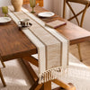 A rustic wooden table is decorated with a beige macrame tassel table runner and simple tableware.