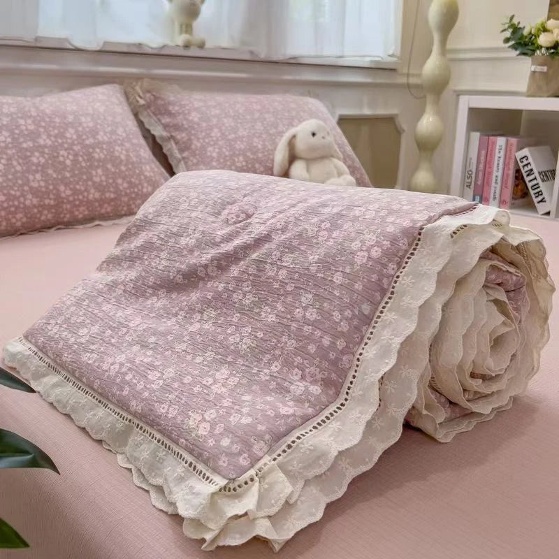 A rolled quilt blanket with a light purple floral print and white lace trim resting on a pink surface.