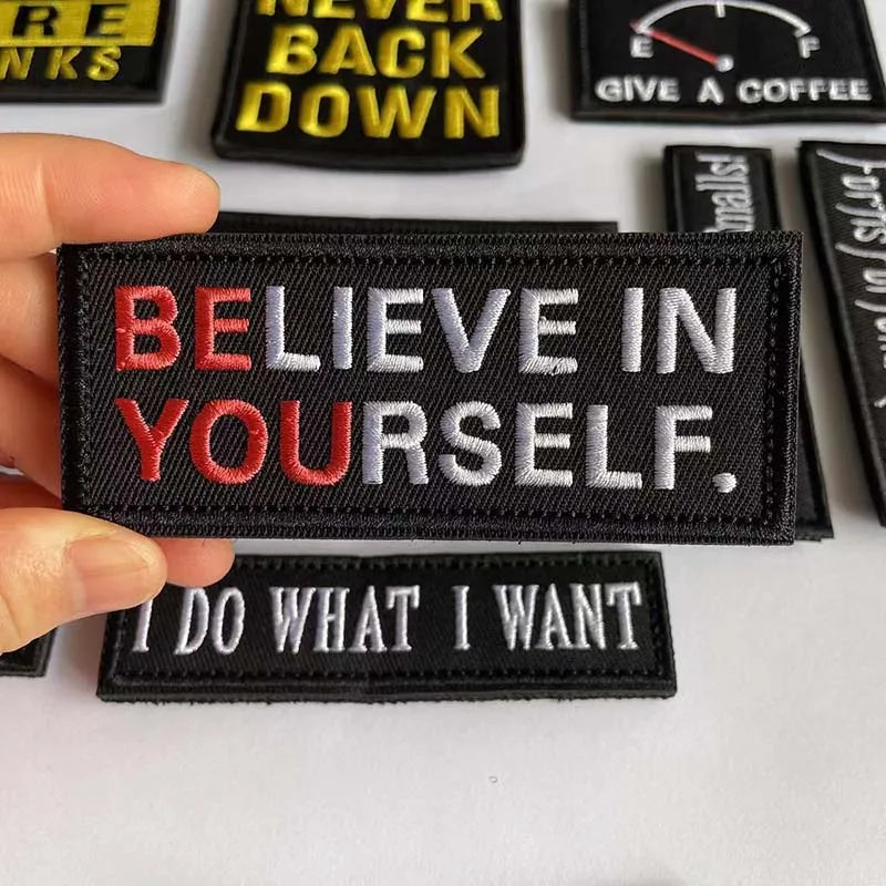 A hand holds several black embroidered alphabet hook and loop patches with motivational text.