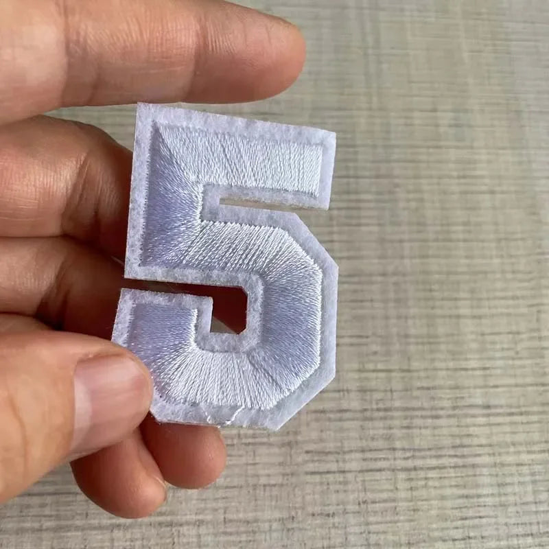 A hand holds a white embroidered number 5 patch for apparel.