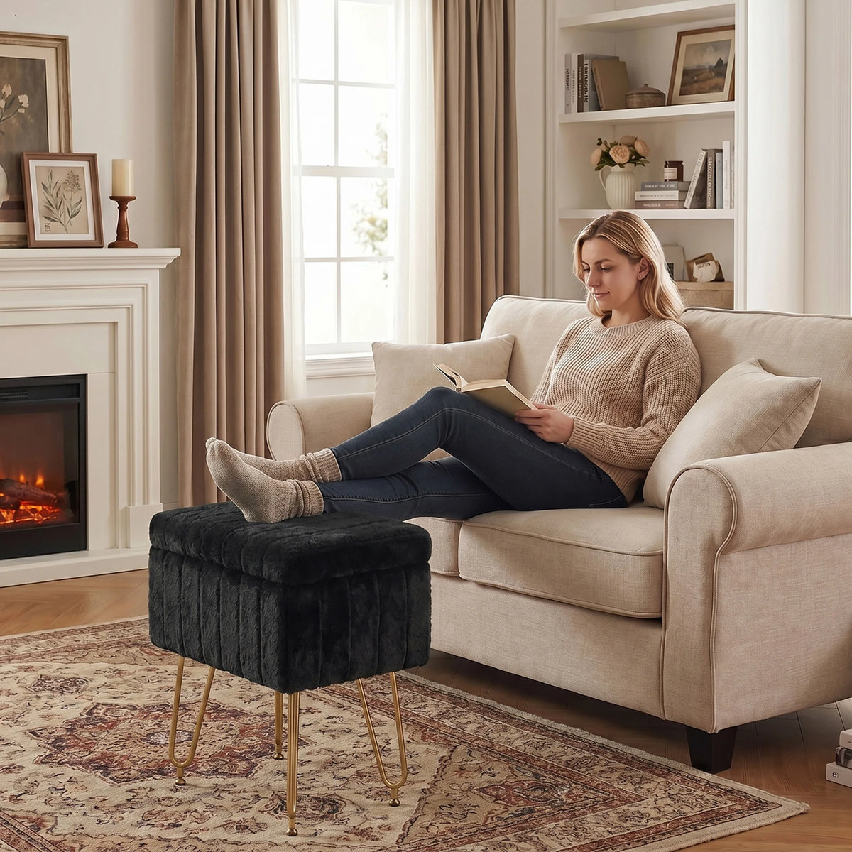 A woman sits on a beige sofa reading a book, next to a dark gray faux fur ottoman with gold metal legs.