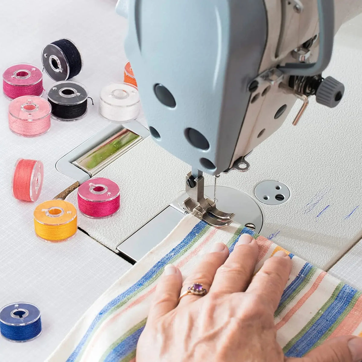 A hand is shown sewing fabric with a clear sewing machine bobbin set and storage case on a white surface.
