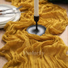 A golden yellow boho crinkle cheesecloth table runner draped over a wooden surface with silverware and a candle holder.