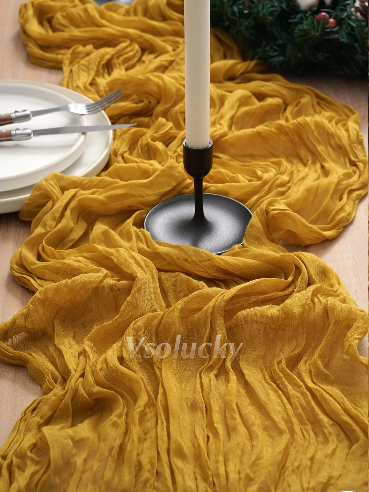 A golden yellow boho crinkle cheesecloth table runner draped over a wooden surface with silverware and a candle holder.