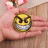 A hand holds a yellow embroidered patch featuring a menacing, grinning face with sharp teeth and angry eyes.